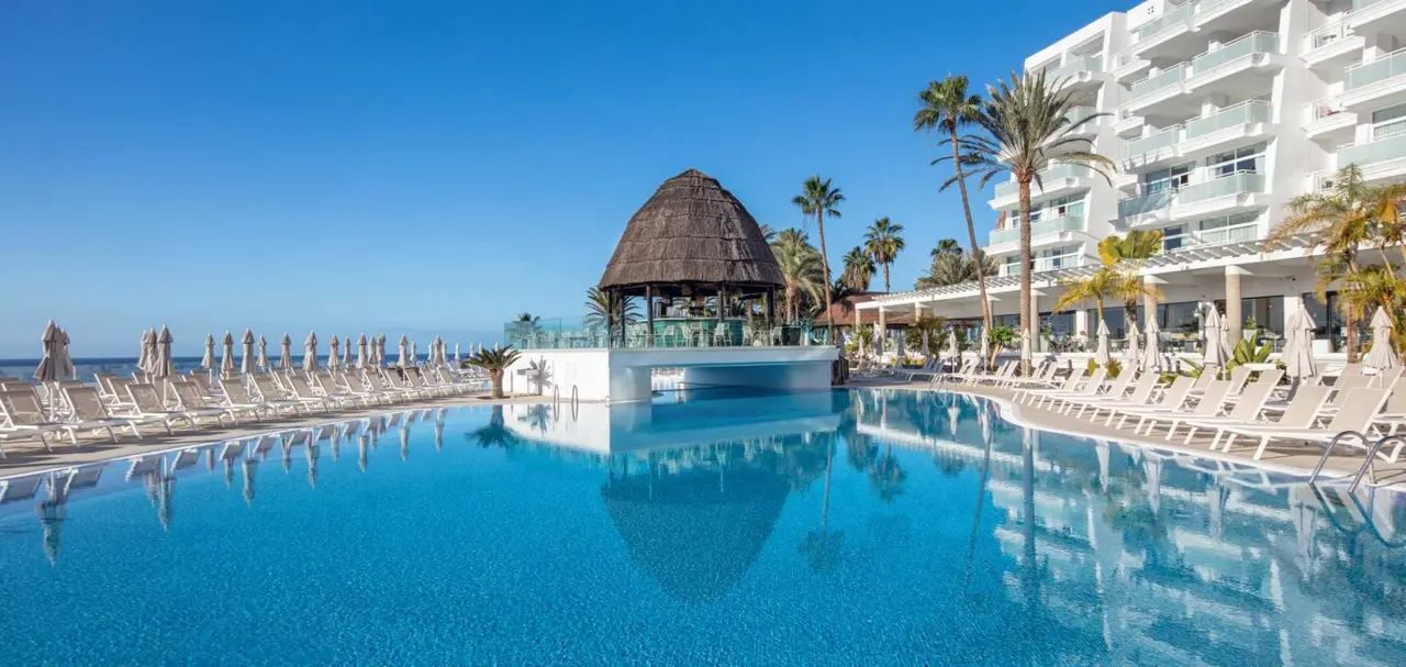 Piscina rectangular con agua azul frente a un palapa y un edificio de apartamentos con palmeras bajo un cielo despejado.