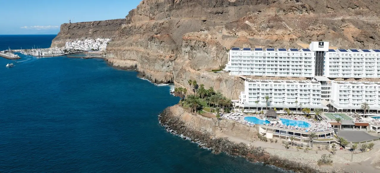 Vista aérea de un hotel blanco junto al mar azul, con piscinas y terrazas al pie de un acantilado rocoso.