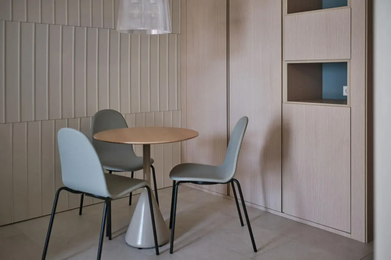 Interior de una suite con mesa redonda de madera y tres sillas modernas de color gris claro con patas negras, situada