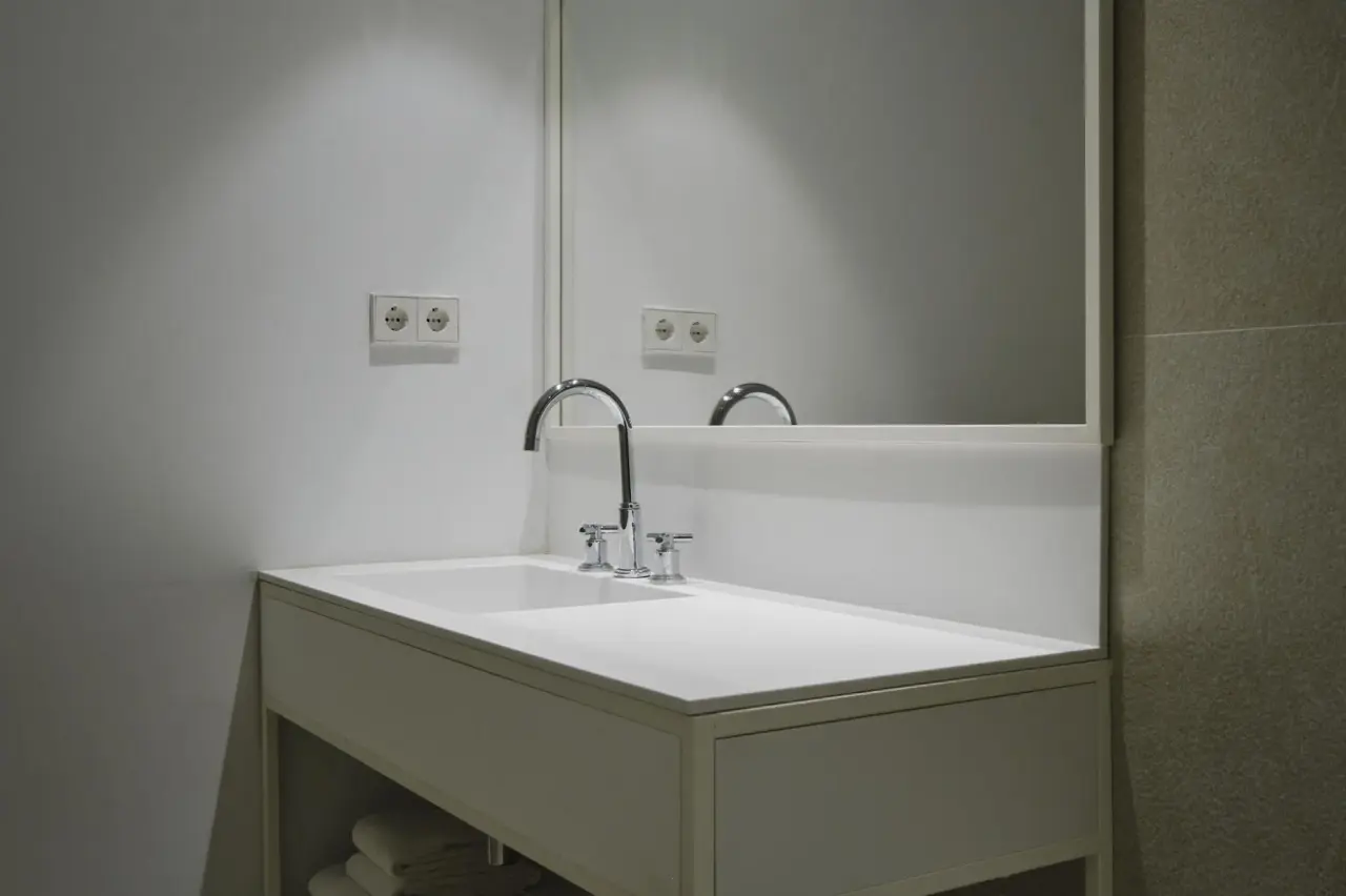 Detalle de un lavabo blanco con grifo cromado, encimera amplia y espejo rectangular sobre él.