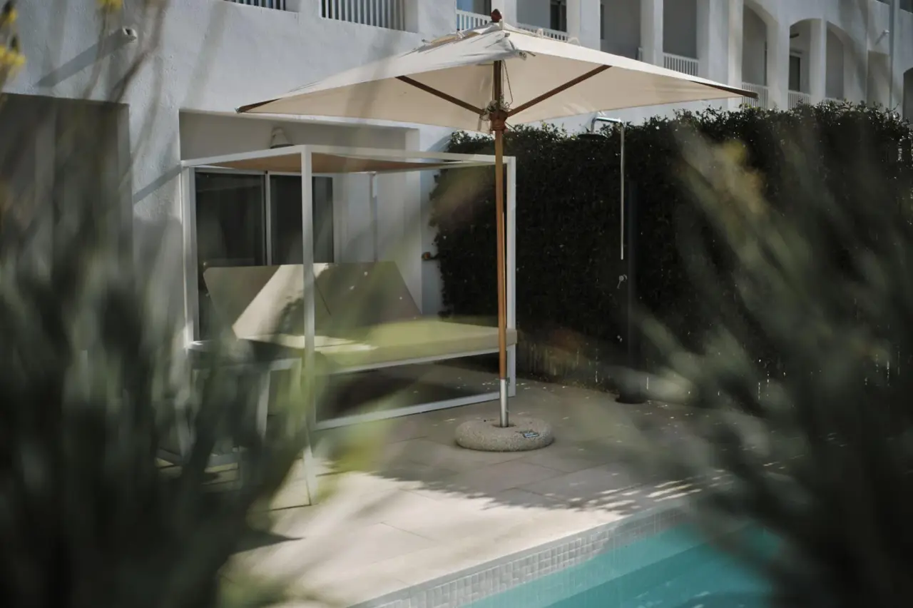 Piscina privada climatizada en una suite, con daybed bajo un parasol, suelo de piedra y edificio blanco al fondo.