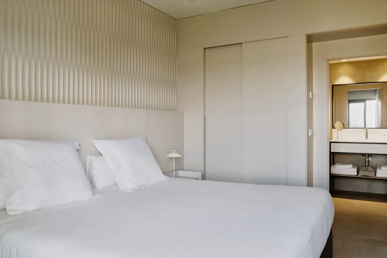 Habitación con cama grande vestida de blanco, cabecero con paneles verticales, mesita de noche con lámpara, armario