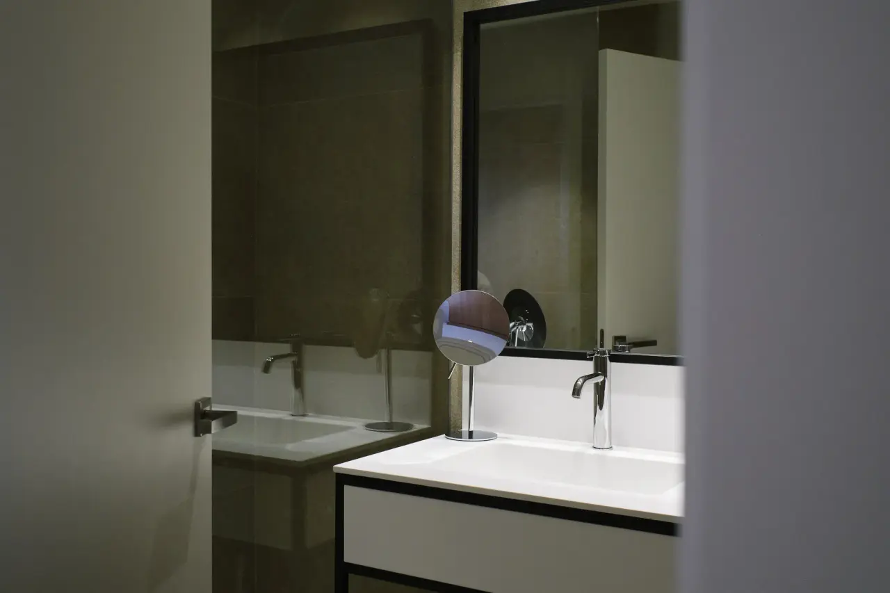 Baño con lavabo blanco rectangular, grifo cromado, espejo grande con marco negro y un espejo de aumento circular