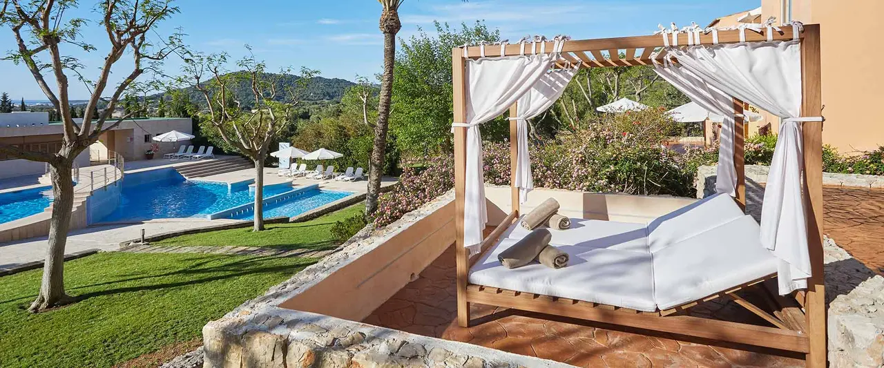 Vista exterior de una terraza con una cama balinesa de madera con dosel y cortinas blancas, sobre un suelo de terracota.