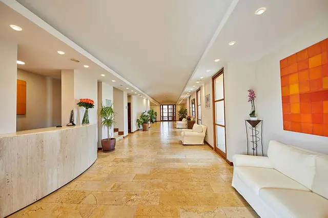 Pasillo interior de un hotel con recepción a la izquierda, sofás blancos a la derecha, suelo de piedra, plantas
