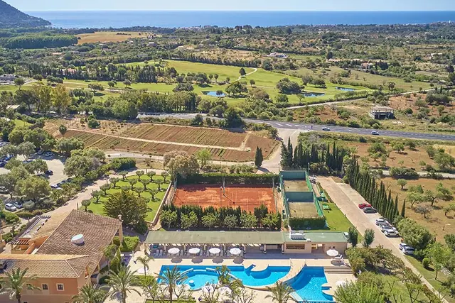 Vista aérea del resort con piscina en primer plano, canchas de tenis, edificios y extenso paisaje rural con mar
