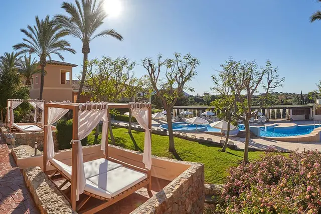 Vista exterior de un resort con varias piscinas, palmeras, césped, árboles y camas balinesas de madera con dosel