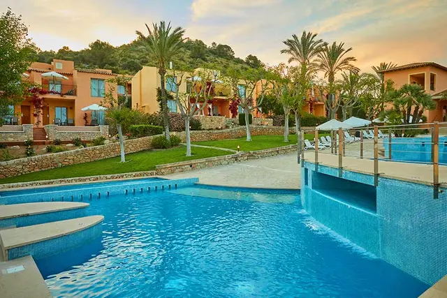 Vista de una piscina rectangular con agua azul y escaleras de acceso en primer plano, rodeada de césped, palmeras