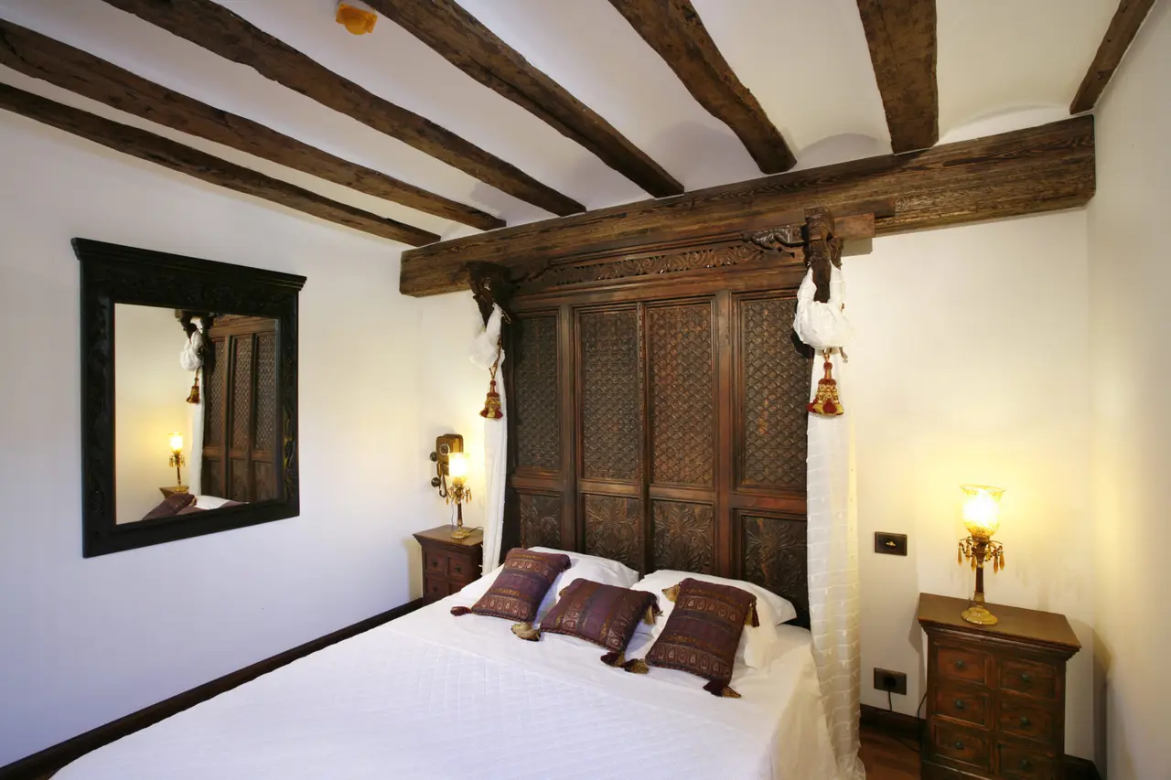 Habitación con cama de dosel de madera tallada, dosel blanco, cabecero ornamentado, mesitas de noche con lámparas