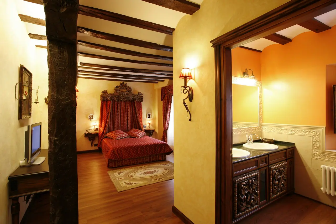 Vista de una habitación de hotel con cama de dosel con cortinas rojas, vigas de madera en el techo, escritorio