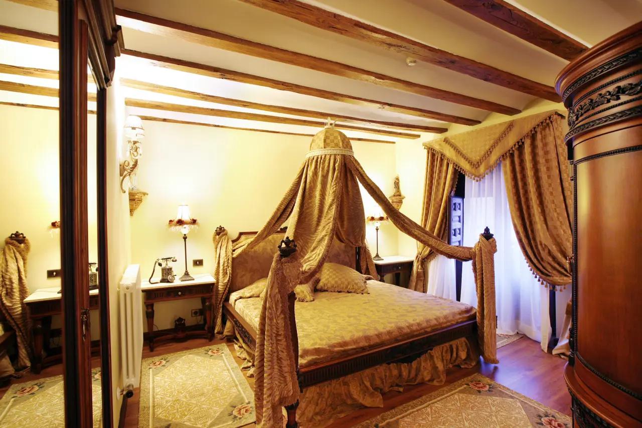 Habitación con cama de dosel grande con dosel dorado, vigas de madera en el techo, cortinas pesadas, mesitas de noche