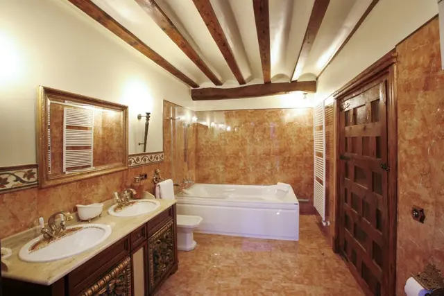 Baño privado con paredes y suelo de mármol rojizo, techo con vigas de madera vistas, doble lavabo sobre mueble