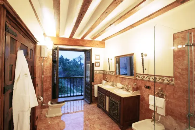 Baño amplio con paredes y suelo de mármol rojizo, techo con vigas de madera vistas, doble lavabo sobre mueble
