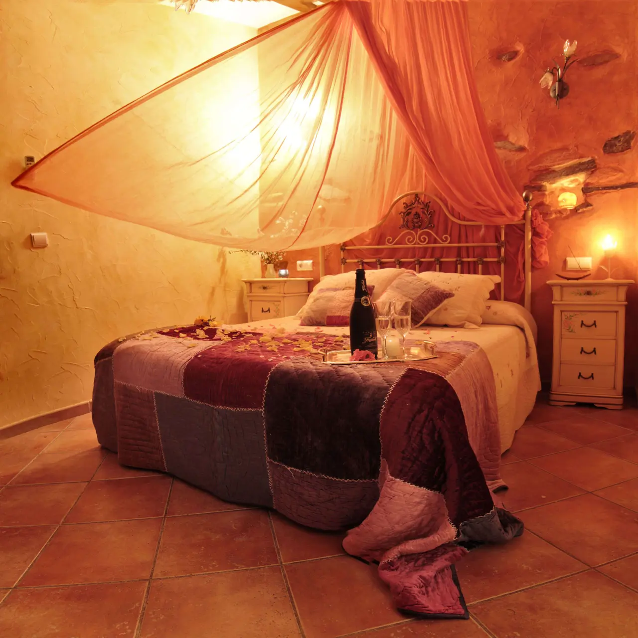 Habitación con cama grande cubierta por un dosel de tela ligera, colcha patchwork en tonos burdeos y grises, mesitas