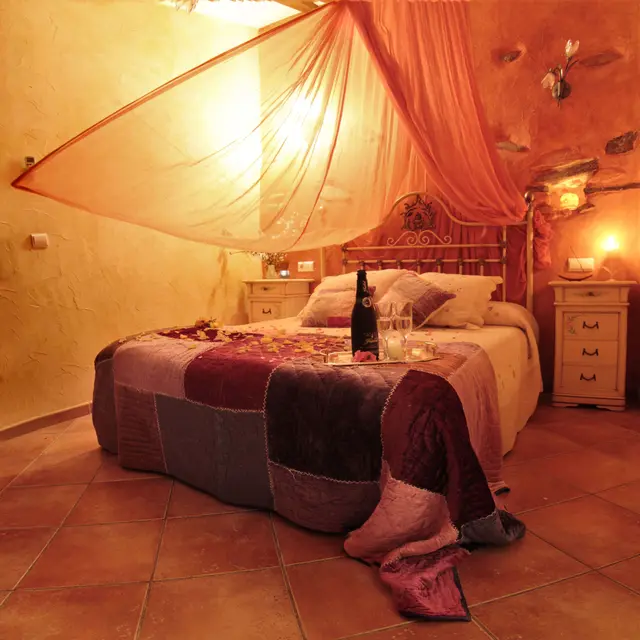 Habitación con cama grande cubierta por un dosel de tela ligera, colcha patchwork en tonos burdeos y grises, mesitas de 