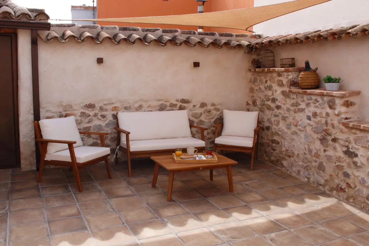 Patio exterior con suelo de baldosas, tres sillones de madera con cojines blancos, mesa de centro de madera