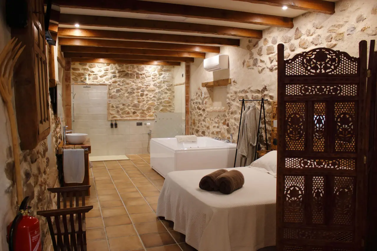 Interior de una habitación rústica con jacuzzi rectangular blanco en el fondo, cama con toallas enrolladas en primer
