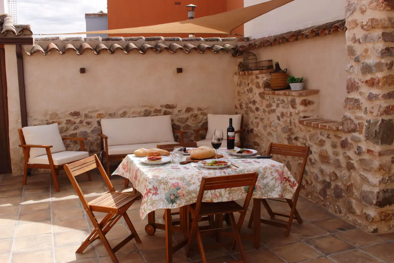 Patio exterior con suelo de terrazo, mesa de comedor redonda con mantel floral y cuatro sillas de madera plegables.