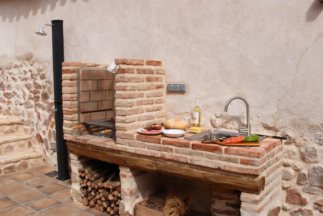 Cocina exterior de obra con encimera de ladrillo, fregadero, parrilla y chimenea de leña adosada a una pared de piedra