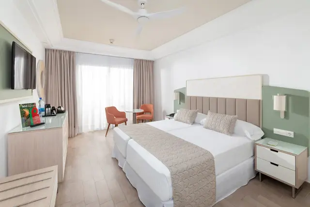Habitación de hotel con cama doble grande vestida de blanco y beige, cabecero acolchado, mesitas de noche, televisor