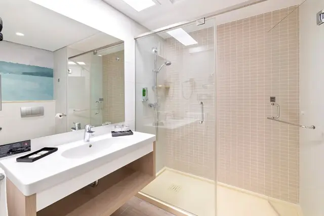 Baño moderno con lavabo blanco sobre encimera de madera, espejo grande, ducha con mampara de vidrio y azulejos beige