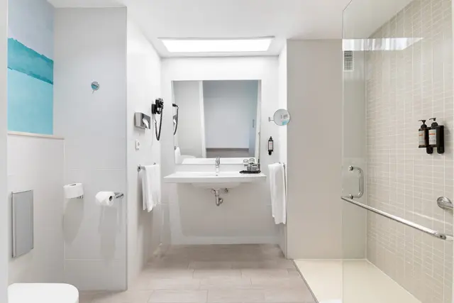 Baño interior de suite con ducha a ras de suelo, lavabo suspendido con espejo, inodoro y toalleros con toallas blancas.