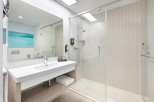 Baño de suite con ducha de cristal, lavabo blanco sobre mueble de madera, gran espejo y toallas dobladas.
