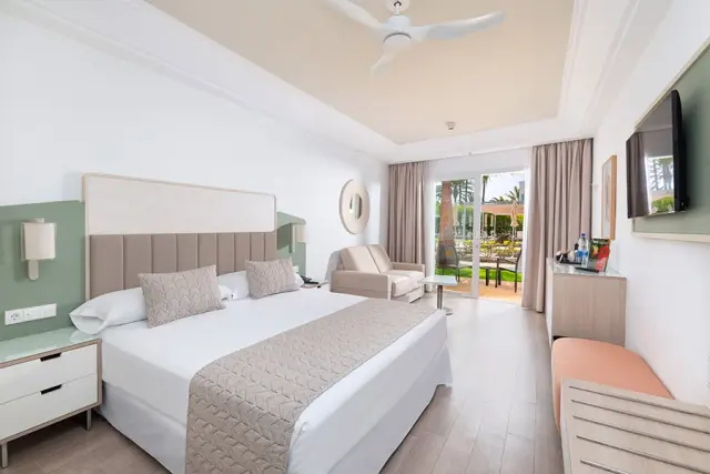 Interior de una suite con cama king size vestida de blanco, cabecero acolchado, dos mesitas de noche, sofá, televisor