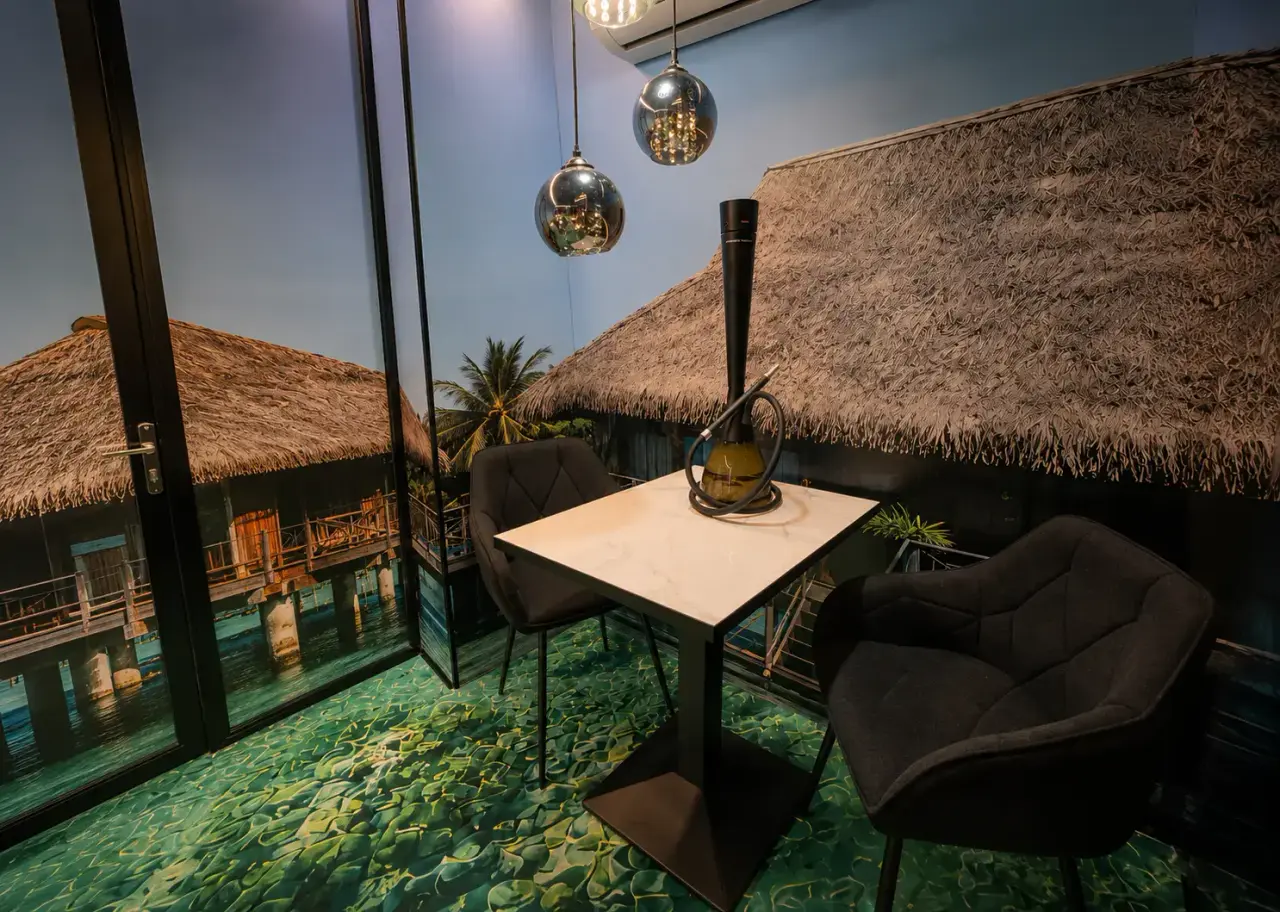 Interior de una habitación con suelo de mosaico verde, mesa blanca con dos sillas oscuras, lámparas colgantes