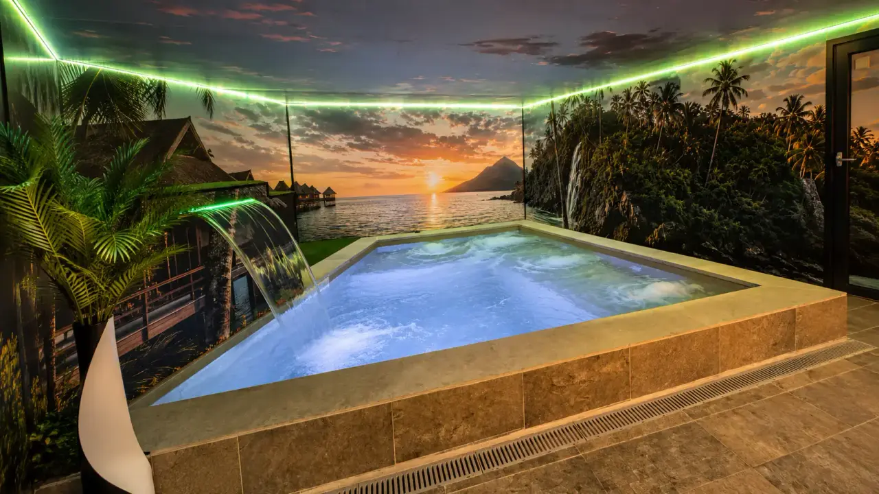 Jacuzzi rectangular interior con agua iluminada en azul, cascada lateral, rodeado de paredes con mural de atardecer