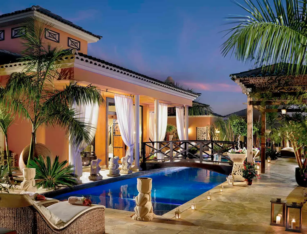 Piscina privada rectangular iluminada al atardecer, rodeada de terraza de piedra, palmeras, tumbonas y una villa