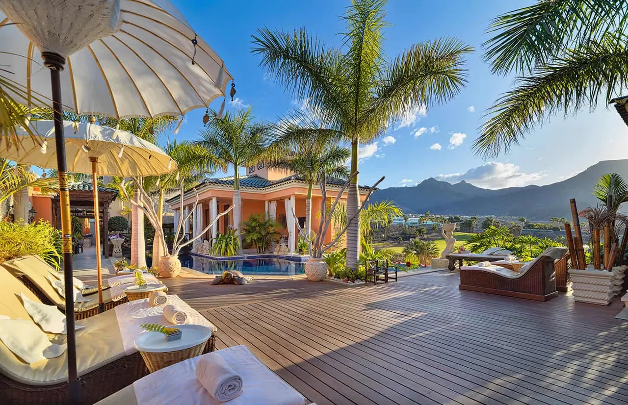 Terraza de madera con piscina privada climatizada, tumbonas, sombrillas y vistas a montañas y palmeras.