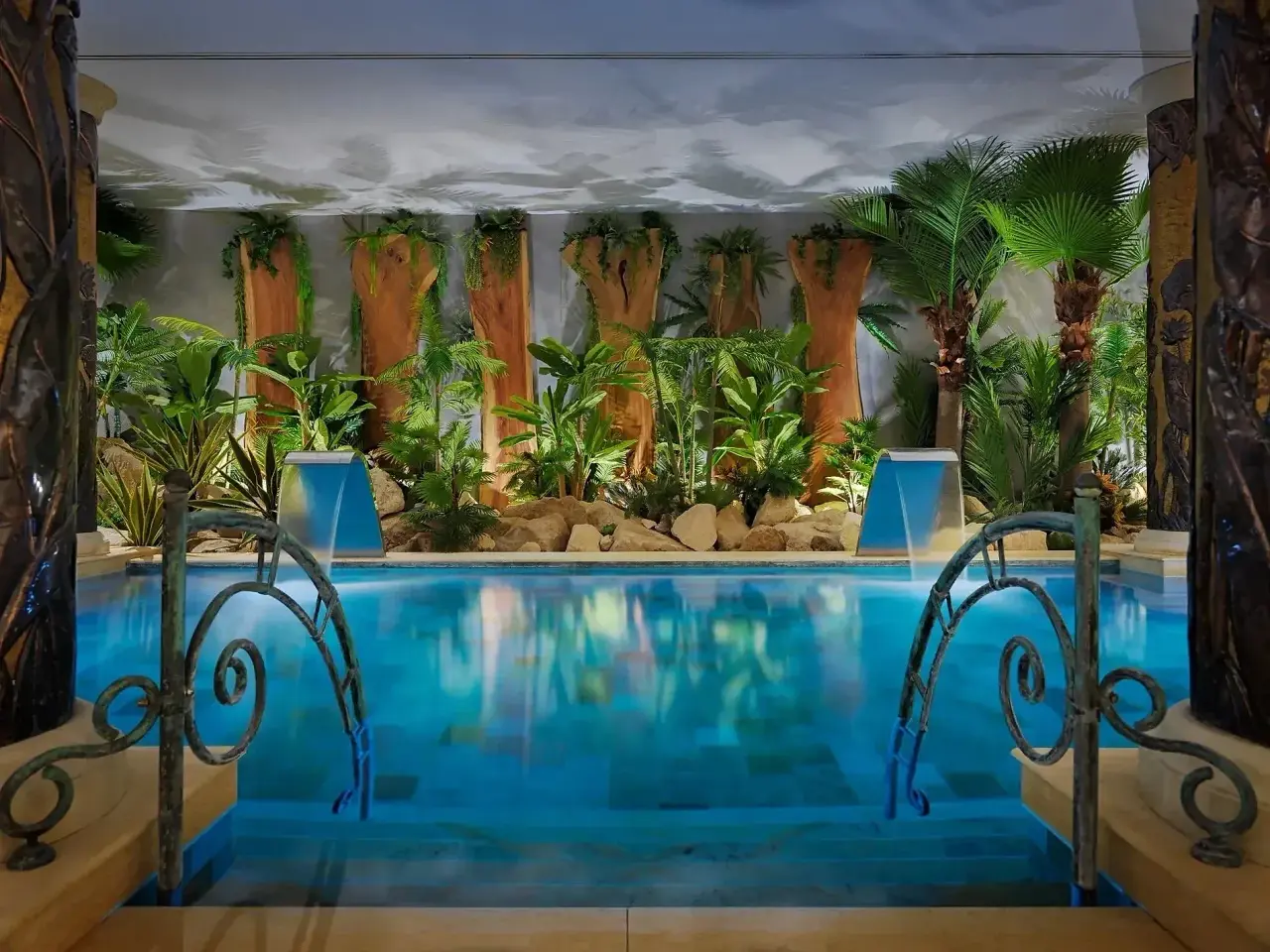 Piscina interior rectangular con agua azul clara, rodeada de vegetación tropical, palmeras y troncos decorativos.