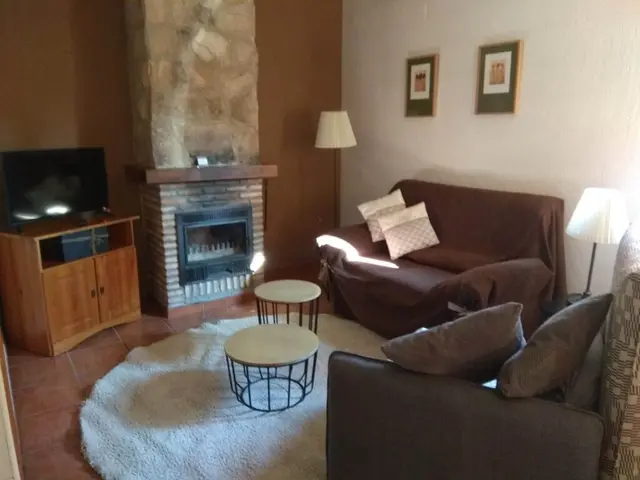 Interior de una sala de estar con chimenea de piedra, televisor sobre un mueble de madera, sofá marrón, sillones, mesa