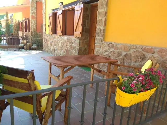 Terraza exterior de una cabaña rural con suelo de baldosas, mesa y sillas de madera con cojines amarillos, barandilla