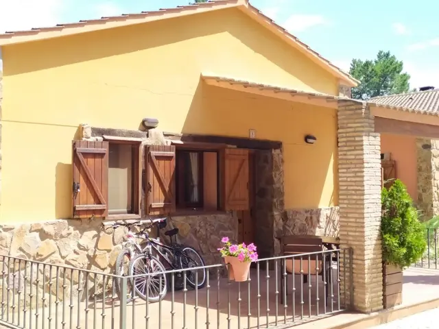 Fachada exterior de una cabaña rural de una planta con paredes amarillas y base de piedra, ventanas con contraventanas