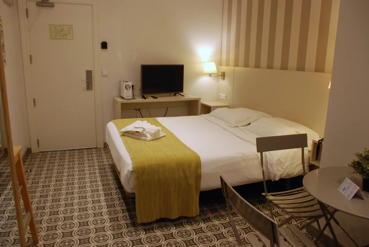 Vista de una habitación doble con cama grande vestida de blanco y una manta amarilla, escritorio con televisor, silla