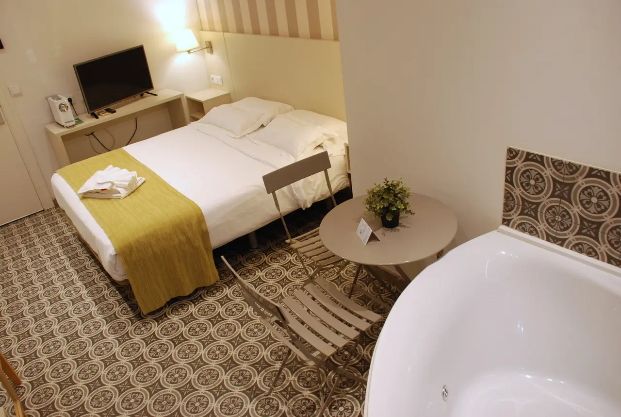 Vista de una habitación de hotel con una cama doble, un jacuzzi blanco en primer plano, una mesa redonda