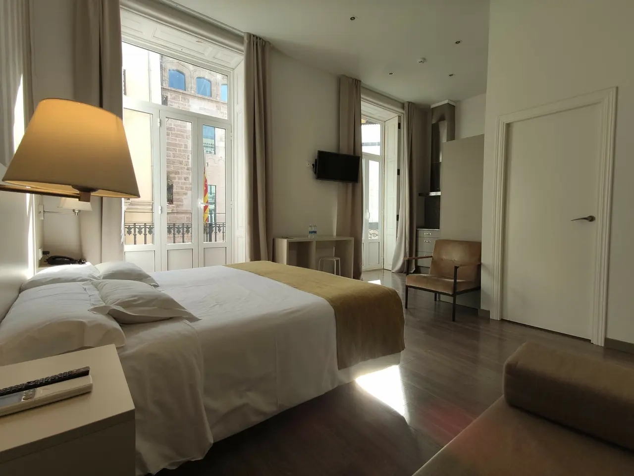 Habitación doble de hotel con cama grande vestida de blanco, lámpara de pie, ventana grande con cortinas, televisión