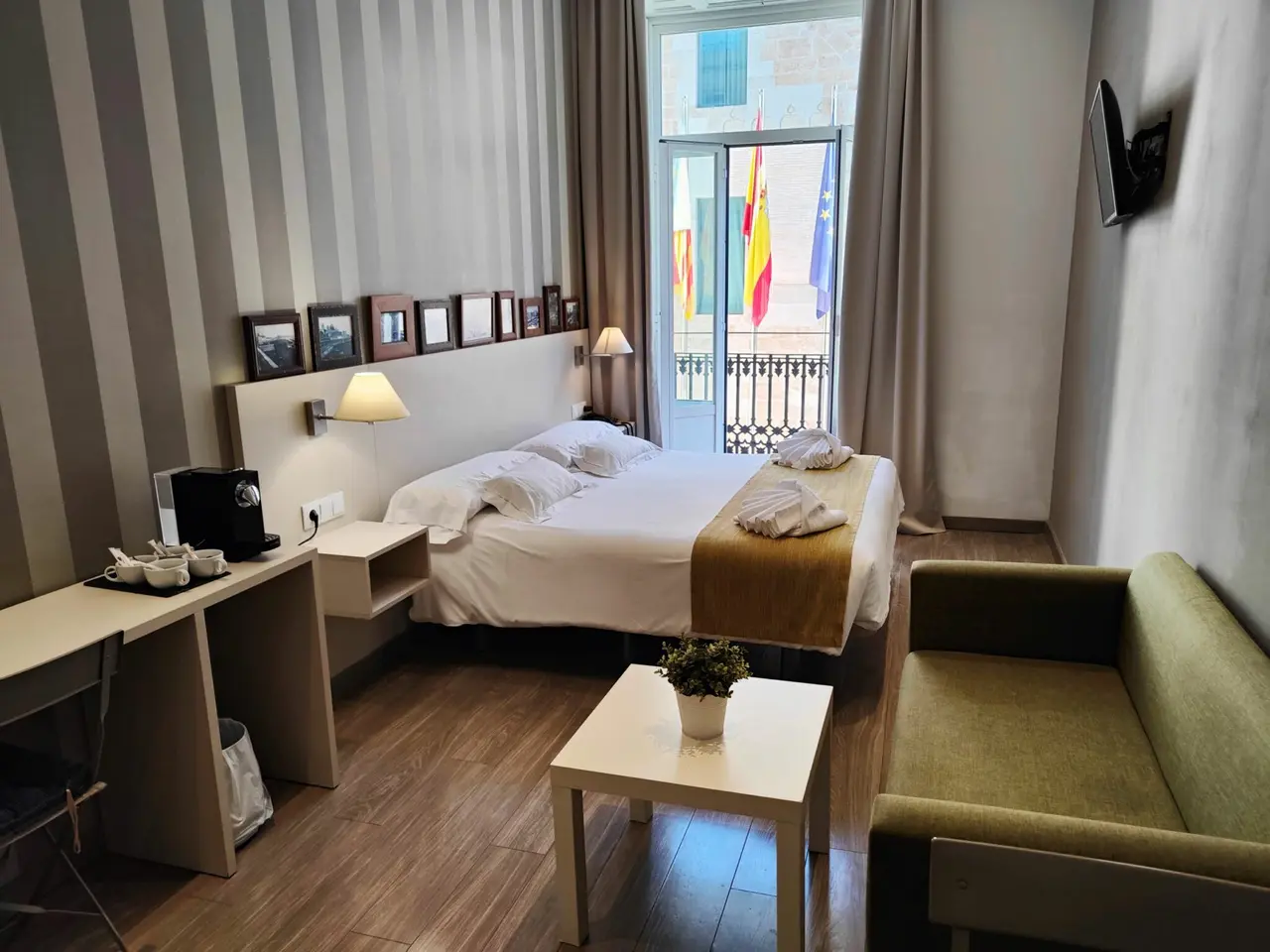 Vista general de una habitación doble con cama grande vestida de blanco, escritorio con máquina de café, sofá verde