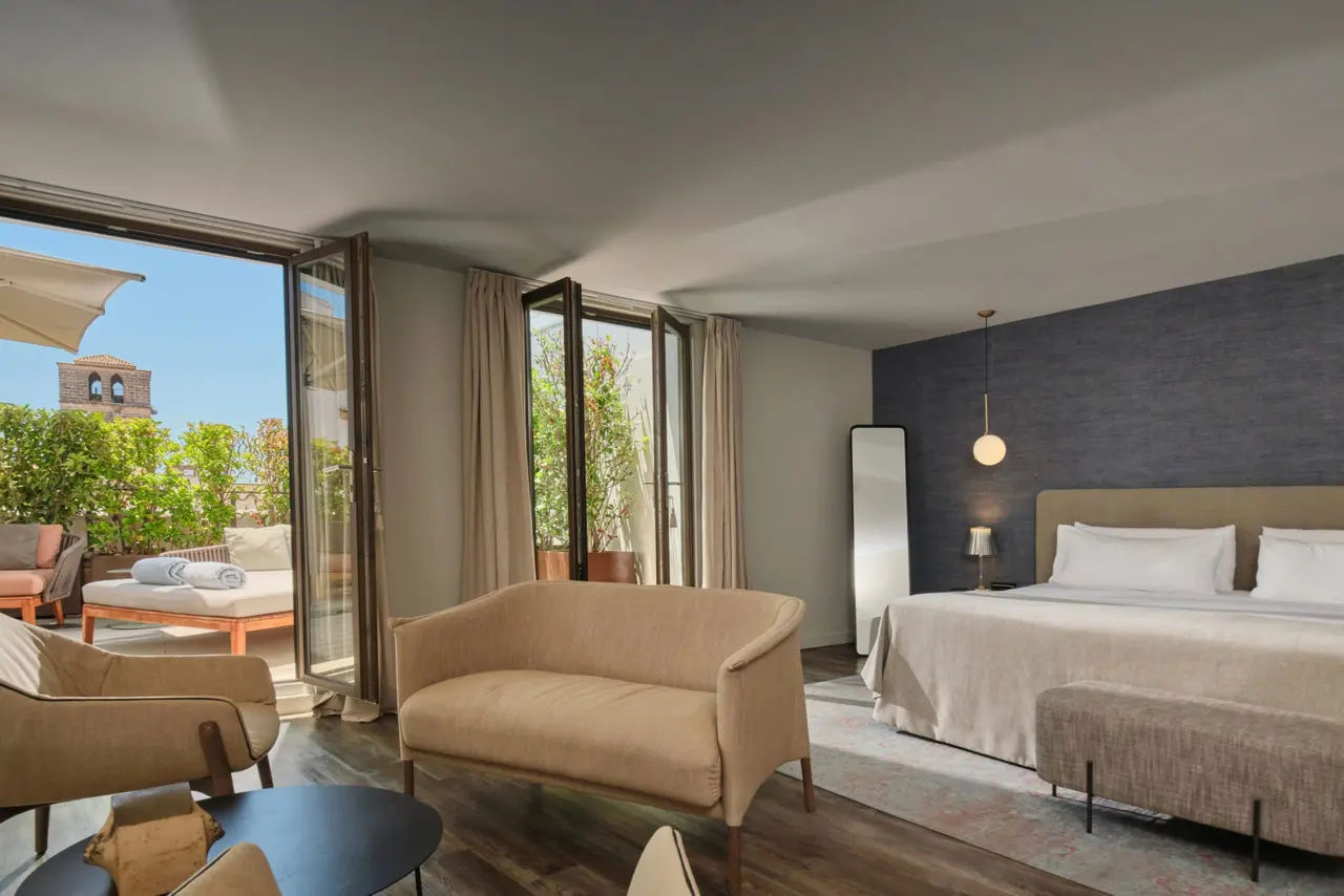 Suite de hotel con cama grande, sofá beige, mesa redonda y puertas abiertas a una terraza soleada con vistas