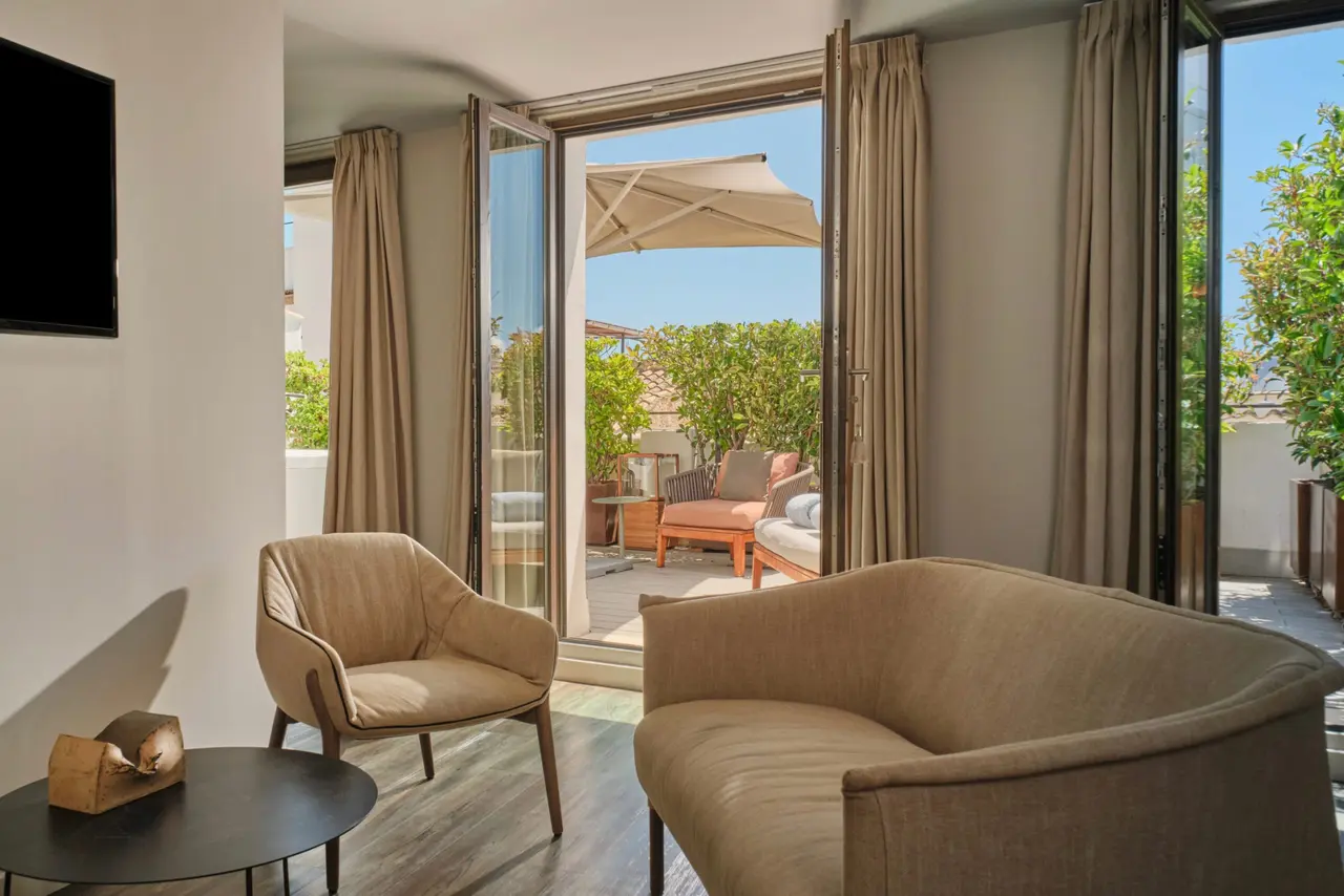 Interior de una suite con sofá y sillón beige, mesa auxiliar y puertas abiertas a una terraza soleada con muebles