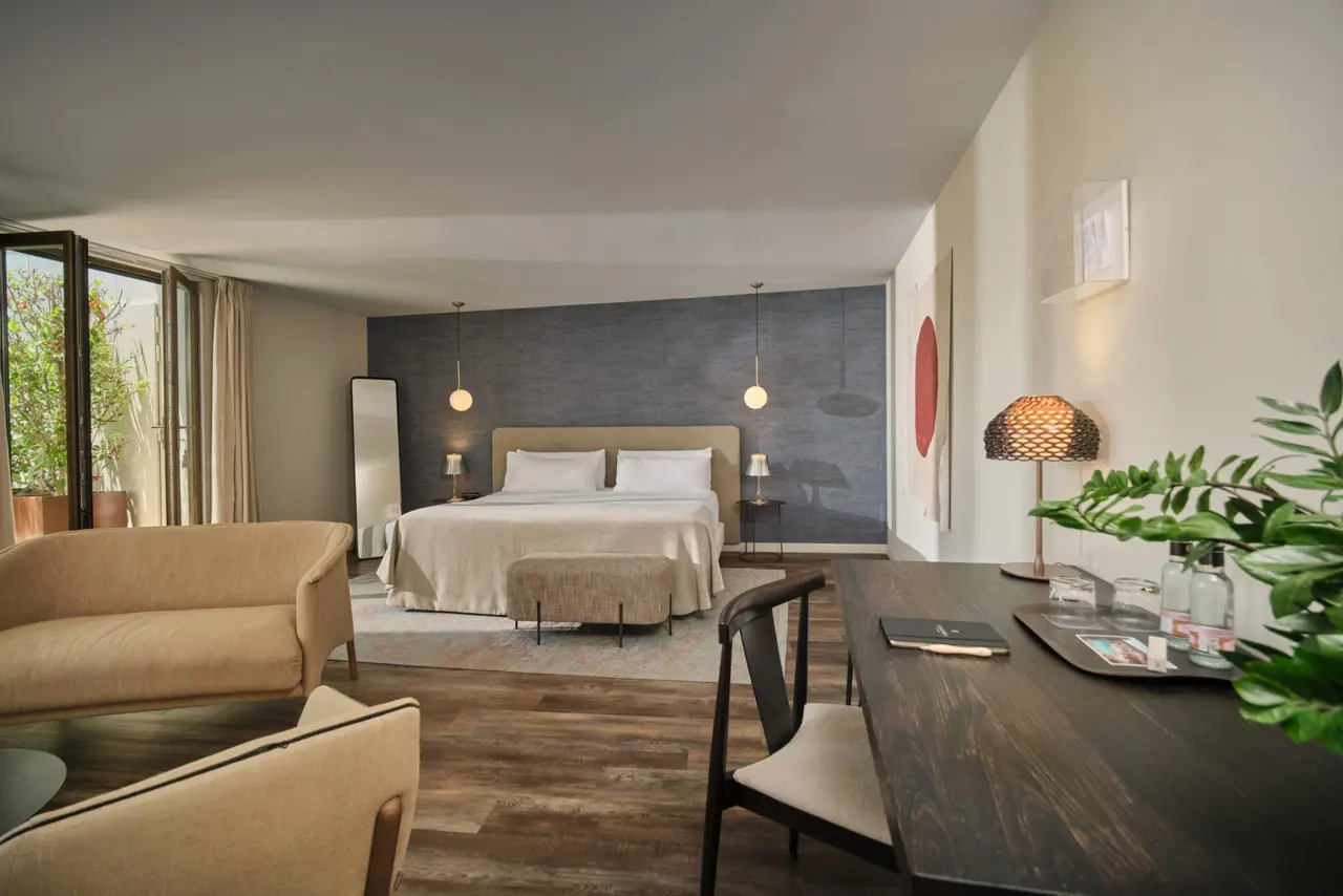 Vista de una suite de hotel con cama grande, sofá, escritorio y puertas abiertas a una terraza exterior.