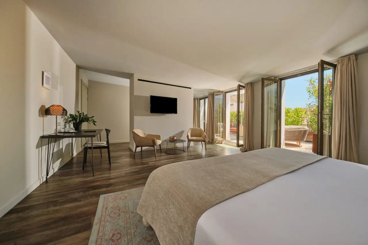 Suite de hotel con cama grande, zona de estar, escritorio, TV y puertas abiertas a una terraza soleada.