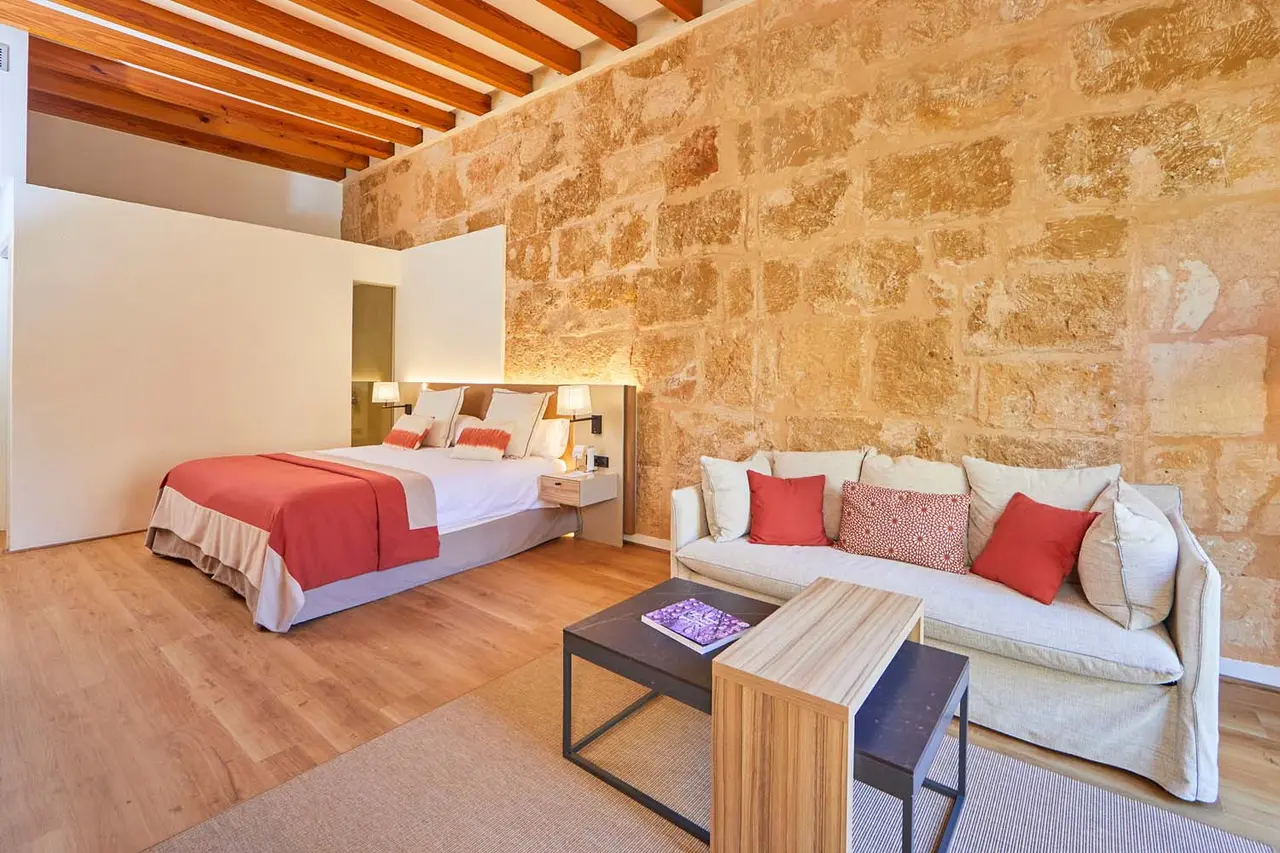 Vista de una suite con cama doble, sofá de tres plazas con cojines rojos, mesa de centro, suelo de madera y pared
