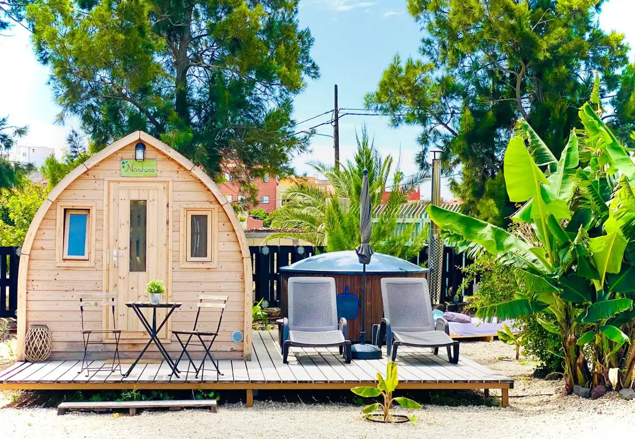 Cabaña de madera con forma de barril sobre una terraza de madera, con mesa y sillas, dos tumbonas y una bañera nórdica