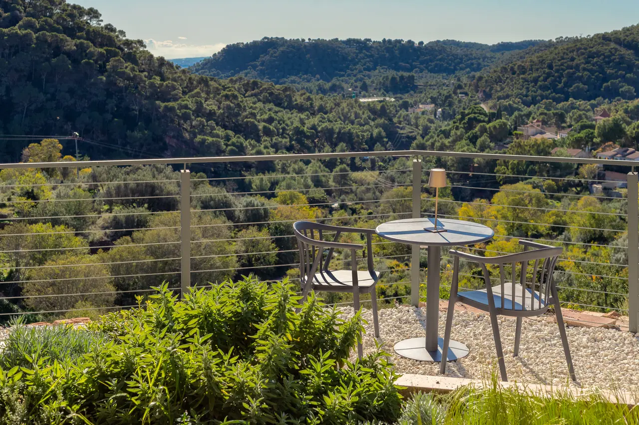 Vista panorámica desde una terraza con mesa y sillas hacia un valle boscoso con colinas cubiertas de vegetación bajo