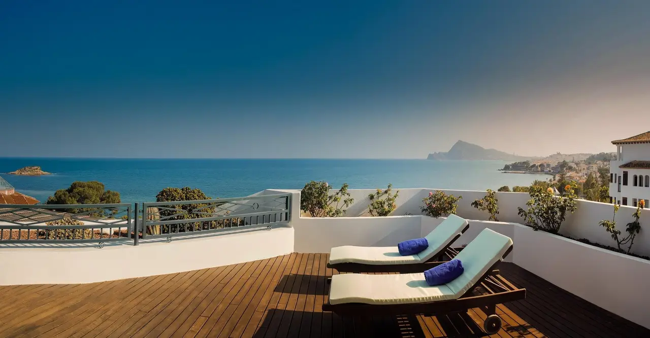 Terraza privada con suelo de madera, dos tumbonas con toallas azules y vistas al mar y la costa.