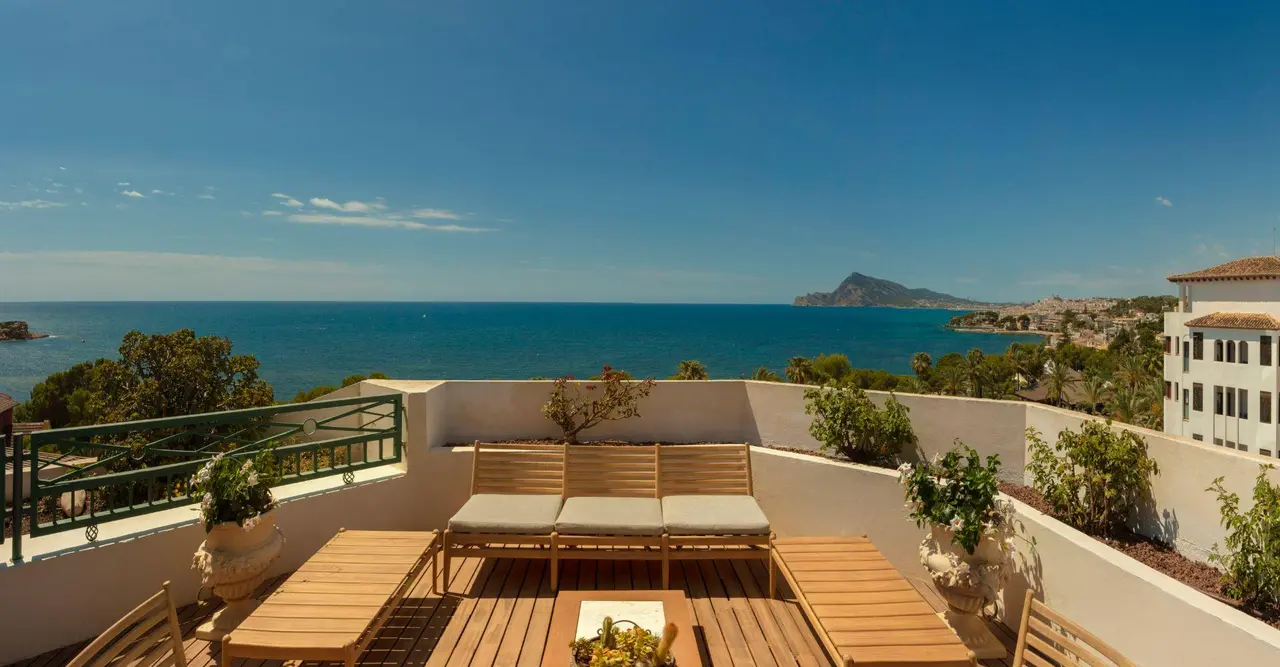 Terraza con suelo de madera, sofá y tumbonas de exterior, barandilla verde y macetas con plantas, con vistas al mar