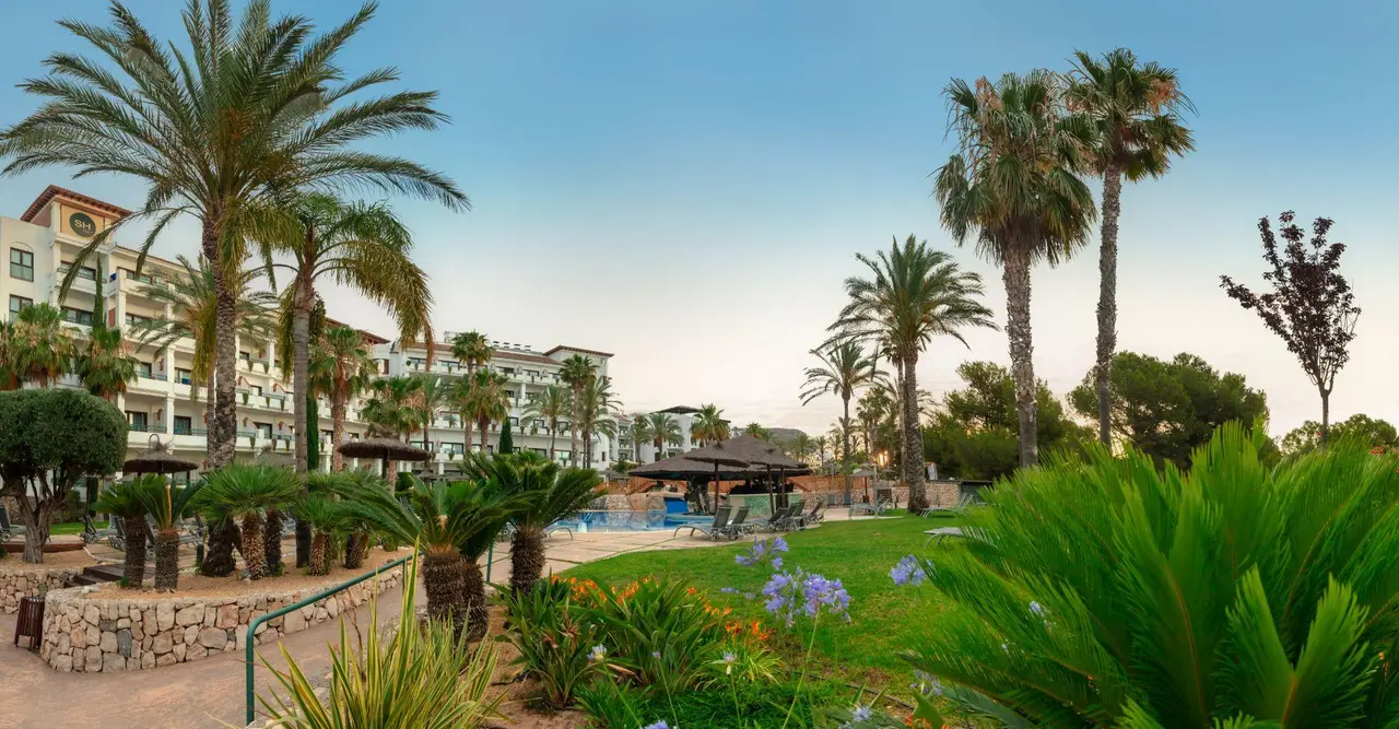Vista exterior de un resort con edificios de alojamiento al fondo, palmeras, jardines con plantas y flores, césped
